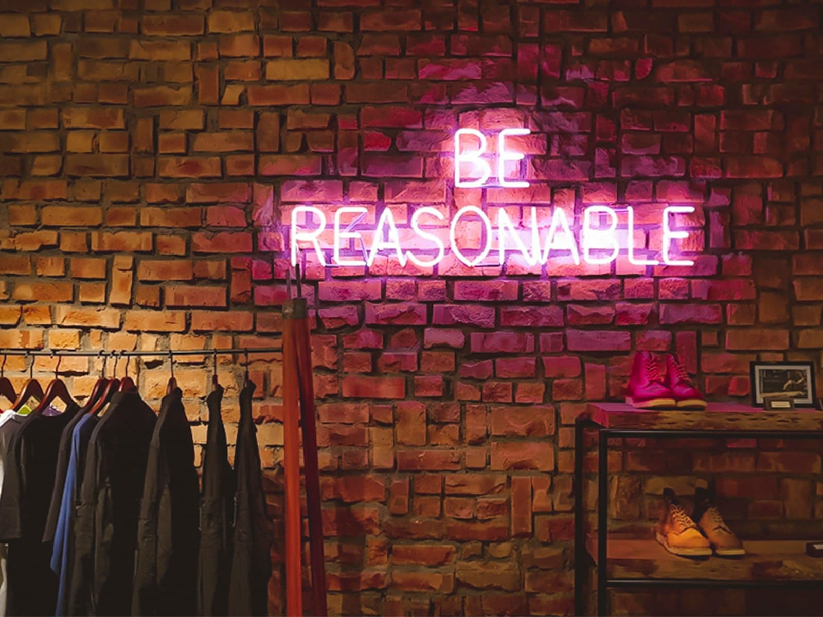 Pink neon sign reading "Be reasonable" on a brick wall